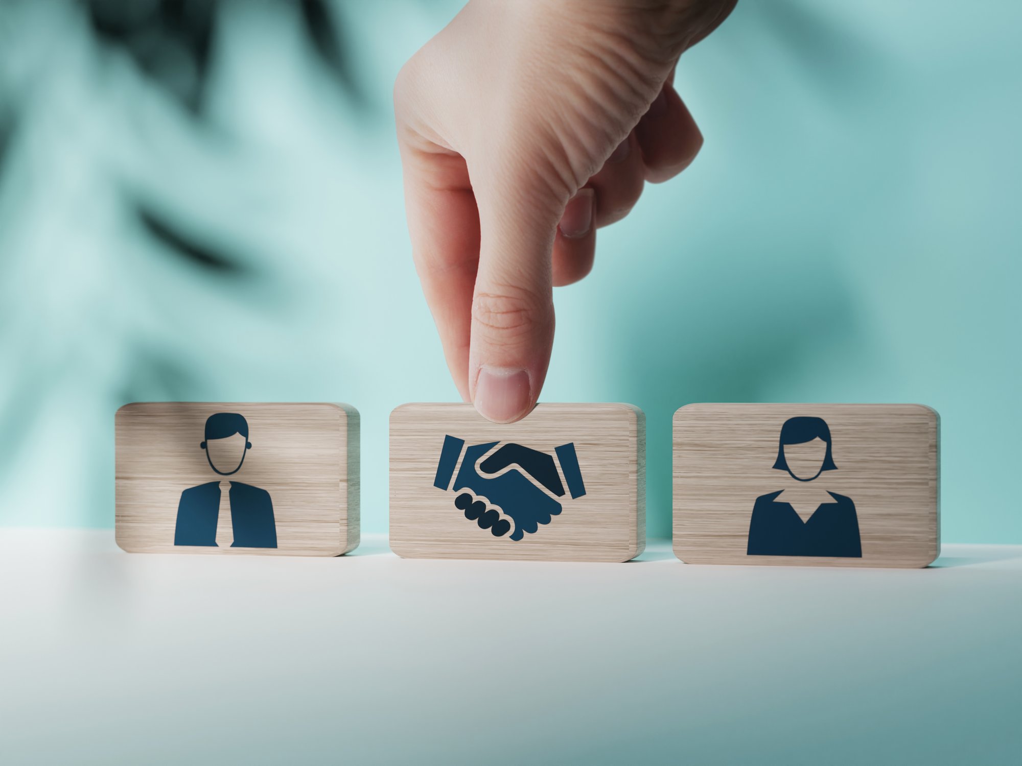 From left to right a wooden tile with a photo of a man, a tile with a photo of hands shaking, and a tile with a woman sit on a white table. A hand reaches down to grab the middle tile with the photo of hands shaking.